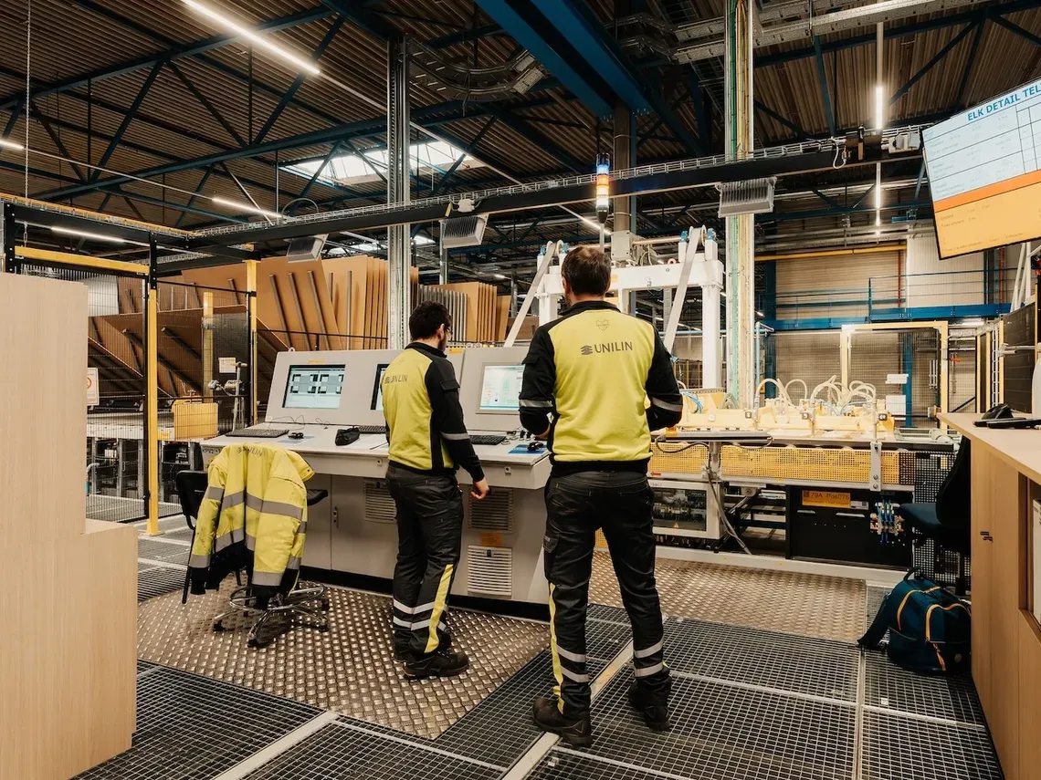 Twee operatoren in gele Unilin-hesjes bedienen de schermen van een controlepost middenin de fabriekshal
